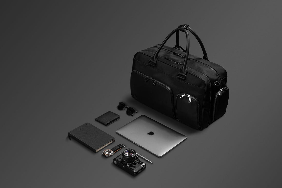 The black Kennedy Weekender with a laptop, notebook, camera, wallet and travel gear laid out beside it